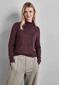 Pull à col roulé bordeaux foncé en maille douce, associé à un pantalon beige clair plissé avec des poches latérales.