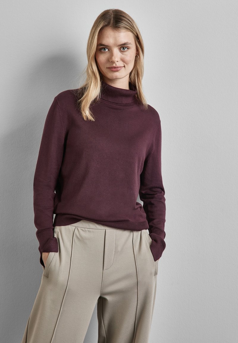 Pull à col roulé bordeaux foncé en maille douce, associé à un pantalon beige clair plissé avec des poches latérales.