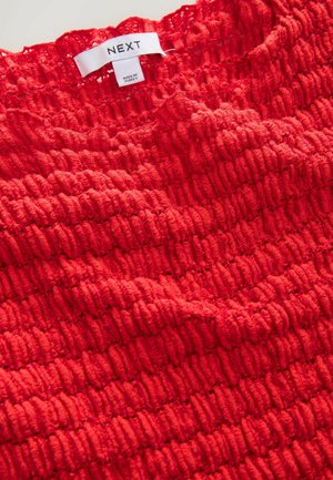 Röd texturerad topp med en smockad design, gjord av mjukt tyg med ett ribbat mönster, som visar en etikett med "NEXT"-logotyp.