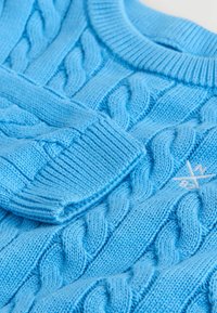 Svetlo modrý pletený sveter s rebrovaným okrúhlym výstrihom, ktorý má tkanú textúru a malú logo na prednej strane.