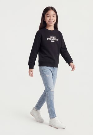 Fiatal lány mosolyogva sétál, fekete pulóvert visel fehér felirattal, világoskék szakadt farmerral és fehér tornacipővel, egyszínű háttér előtt.