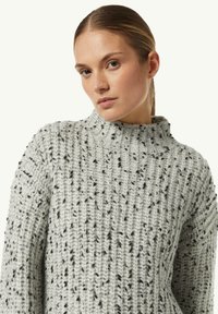 Helles graues Strickpullover mit hohem Kragen, grober Textur und schwarzen Sprenkeln über das gesamte Kleidungsstück.