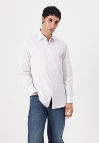 Bela srajca z dolgimi rokavi in gumbi iz gladkega materiala, z klasičnim ovratnikom, v kompletu z modrimi jeans hlačami.