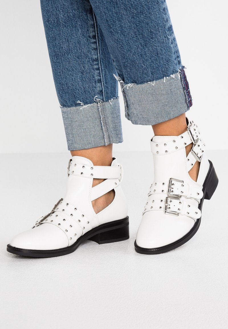 Bottines blanches avec clous et boucles argentées portées avec un jean bleu retroussé sur fond blanc.