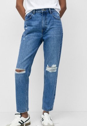 Person wearing blue high-waist ripped jeans with holes on knees, white sneakers with black stripes, and white shirt tucked in.