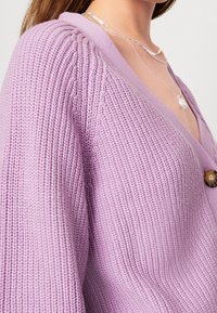 Lavendelstickad kofta med V-ringning, texturerad ribbning och rund brun knappknäppning. Accentuerad med delikata silversmycken.