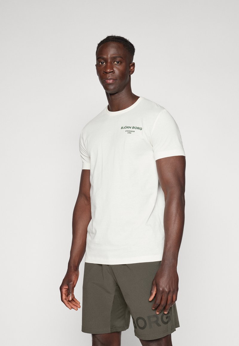 Tricou alb din bumbac, cu guler rotund și logo verde pe pieptul stâng. Purtați cu șorturi verzi măslinii, având text negru.