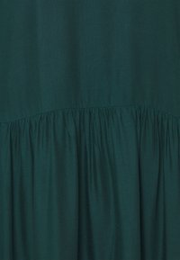 Tissu vert foncé avec un détail de couture froncée, présentant une texture lisse et une silhouette fluide. Le matériau semble léger et respirant.