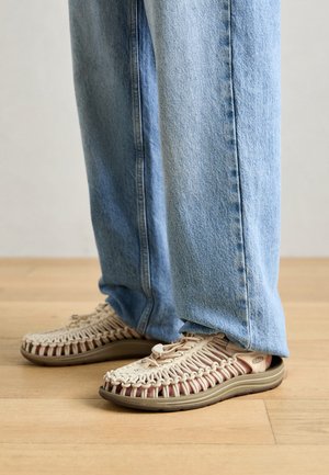 Šviesios spalvos šlepetės su austa viršutine dalimi, turinčios masyvų pado, kuris yra smėlio spalvos ir raudonų akcentų. Priekyje matoma užsegimo sistema su užtrauktuku.