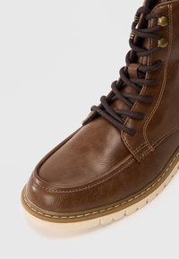 Braune Leder-Schnürstiefel mit runder Zehenform, beiger Gummisohle und kontrastierenden Nähten. Verfügt über Metallösen und eine strukturierte Oberfläche.