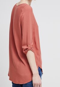 Korallenbluse mit lockerer Passform, Dreiviertelärmeln und Knopfdetails. Glattes Material mit Rundhalsausschnitt und seitlichen Schlitzen am Saum.
