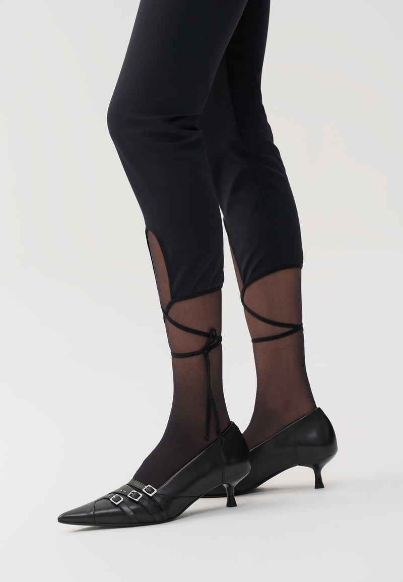 Zwarte kittenheelschoenen met spitse neuzen, voorzien van drie decoratieve gespen. Draagt bij zwarte leggings met doorzichtige panelen en enkelbandjes.