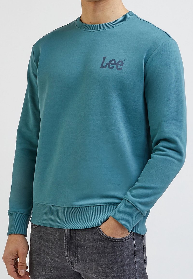 Sudadera turquesa con cuello redondo y mangas largas. Presenta un pequeño logo "Lee" en azul oscuro en el pecho izquierdo y puños y dobladillo acanalados.