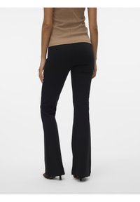 Pantalon noir évasé en tissu extensible, présentant une texture lisse et des coutures discrètes, associé à des chaussures à talons hauts marron.