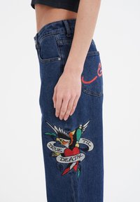 Denim jeans with a high waist, featuring colorful embroidery of a heart, bird, and banner reading "TRUE TIL DEATH" on the leg.