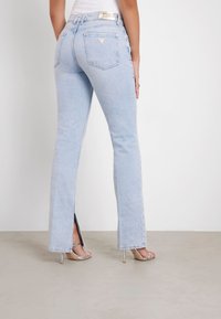 Jeans de color azul claro con un ligero flare, que presentan aberturas laterales, dos bolsillos traseros y una etiqueta de marca en la cinturilla. Textura de denin suave.