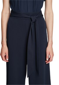Sötét tengerészkék, bő szabású nadrág, a derékrésznél megkötött szövetövével. Sima anyagból készült, tiszta és strukturált sziluettel.