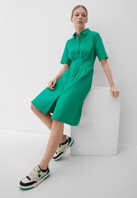 Zielona sukienka koszulowa z lekkiego materiału, z kołnierzykiem, krótkimi rękawami i zapięciem na guziki z przodu. Połączona z kremowo-czarnymi trampkami z zielonymi akcentami.