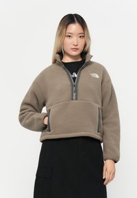Beige fleecegenser med høy krage, halv glidelås og kontrasterende sorte glidelåser. Myk tekstur, to sidelommer og logo på brystet.
