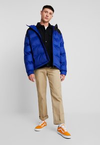 Blue puffer jacket with elastic cuffs, paired with a black shirt, khaki pants, and orange sneakers. Features a zip closure and side pockets.