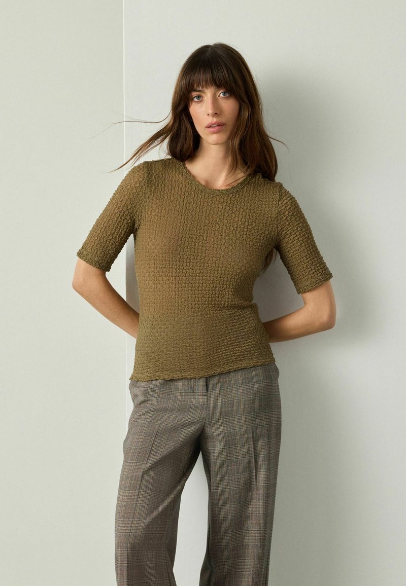 Olivgrünes T-Shirt mit Textur, kurzen Ärmeln und rundem Ausschnitt, kombiniert mit grauen Karohosen in lässiger Passform.