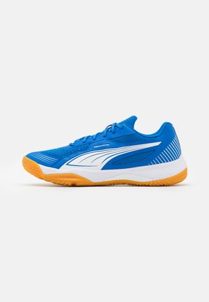 Chaussure de sport d’intérieur bleue à lacets, avec un logo blanc sur le côté, une semelle intermédiaire blanche et une semelle extérieure en gomme orange, représentée de profil sur un fond uni.