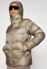 Persoon in een glanzende beige pufferjack met capuchon op, handen in de zakken, en grote zwarte zonnebril tegen een neutrale achtergrond.