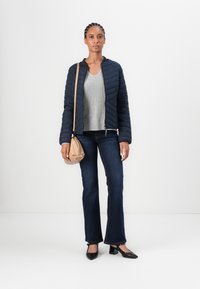 Navy quiltet jakke, grå v-hals genser, mørkeblå utsvingte jeans, svarte blokk-hæl sko og en lysebrun crossover-veske.
