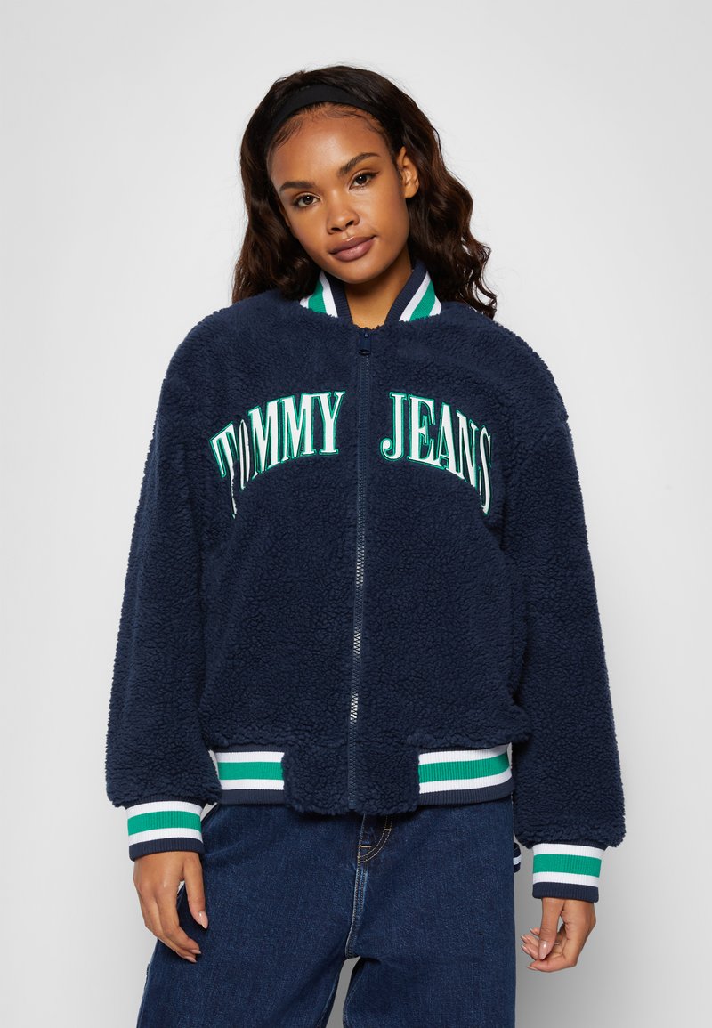 Tumed sinine fliisdžiakk, millel on valged ja rohelised triibulised ribakätised, ning ees on julge kiri "TOMMY JEANS".