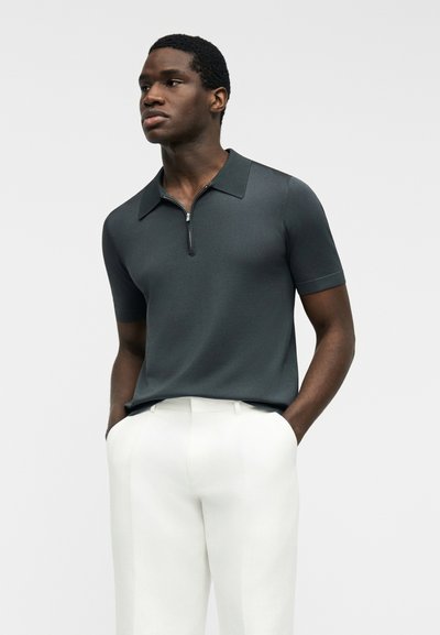 Muž oblečený v tmavě šedé krátkosmetné polo košili na zip a bílých kalhotách, stojící s rukama v kapse proti jednoduchému pozadí.