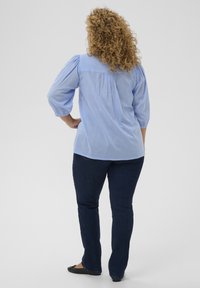 Chemise en coton bleu clair à manches bouffantes et pli au dos, associée à un jean bleu foncé et des chaussures plates noires. Vue de dos.