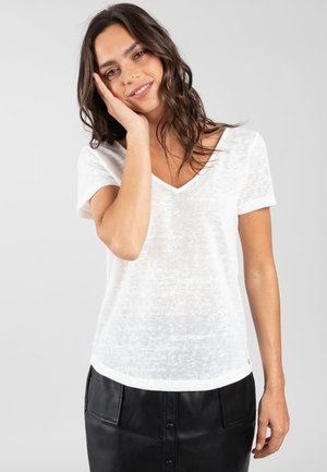 Jeune femme aux cheveux bruns ondulés, souriante, portant un t-shirt blanc à col en V et manches courtes et une jupe noire à boutons, main reposant sur sa joue.