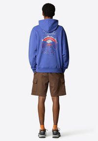 Blue hoodie with a graphic design featuring mountains and figures in motion. Paired with brown cargo shorts. Casual, relaxed fit.