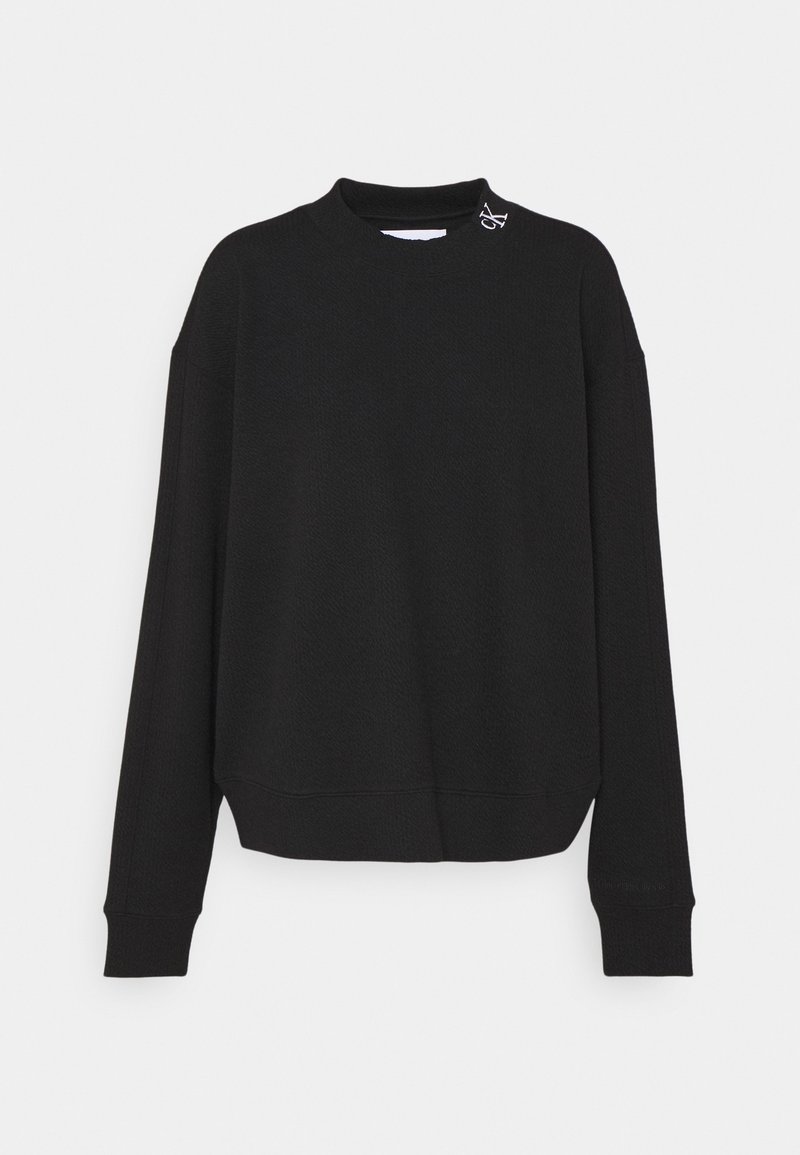 Pull noir à manches longues, col rond, avec un petit logo blanc près de l'encolure côté gauche, présenté sur un fond gris uni.
