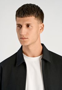 Czarna kurtka na zamek z kołnierzykiem, noszona na białej koszulce. Kurtka ma gładką fakturę i dopasowany krój.