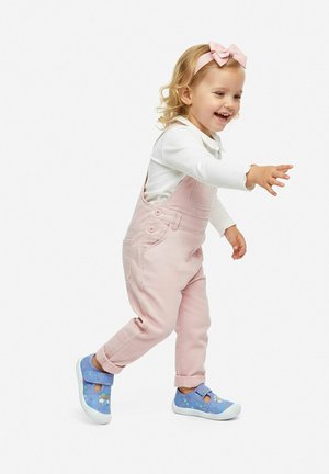 Mała dziewczynka uśmiechająca się i wyciągająca ręce do przodu, ubrana w różowe ogrodniczki, białą koszulkę z długim rękawem, różową opaskę z kokardą oraz niebieskie buty w wzory.