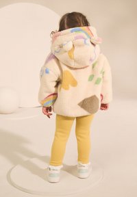 Veste en polaire crème et duveteuse avec une capuche, ornée de motifs multicolores et une pièce marron. Associée à des leggings jaunes et des chaussures blanches.