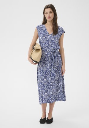 Femme portant une robe midi à motifs bleu et blanc avec un col en V, tenant un chapeau tressé beige et chaussée de chaussures noires, debout sur un fond blanc.
