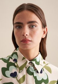 Blusa floreale in morbido tessuto, con grandi fiori verdi e blu, colletto con bottoni e uno sfondo crema.