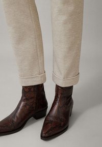 Braune Schlangenhaut-Stiefeletten mit spitzem Zeh und niedrigem Blockabsatz, kombiniert mit gerollten beigen Hosen. Das strukturierte Muster auf den Stiefeletten ist sichtbar.