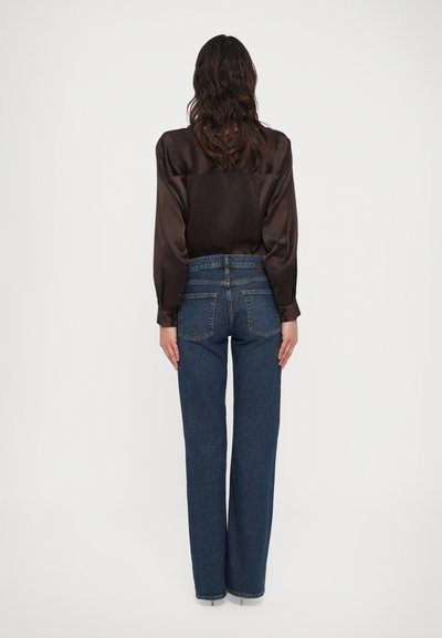 Hosszú sötét hajú nő, háttal áll, sötét szatén hosszú ujjú ingben és magas derekú, kék egyenes szárú farmert visel.