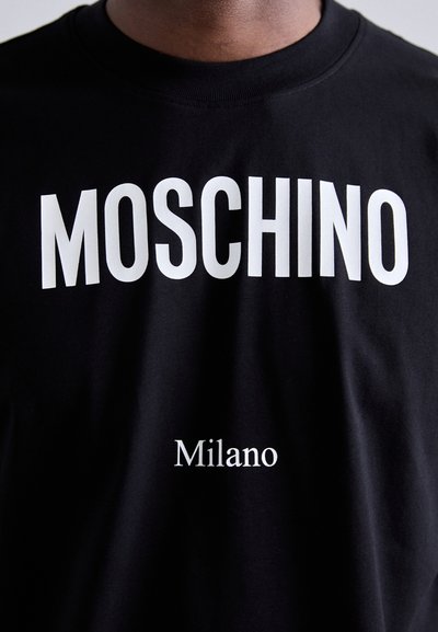 Mustmust T-särk mustast puuvillast, millel on julgelt valged tekstid "MOSCHINO" ülal ja "Milano" all, ümar kaelus ja sile tekstuur.