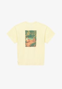 T-shirt jaune clair avec une impression rectangulaire de deux chaises et une table sur un patio à carreaux entouré de plantes tropicales luxuriantes.