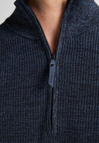 Marineblauwe zip-up trui van textuur gebreide stof, met hoge kraag en een contrasterende rits met een merklabel.