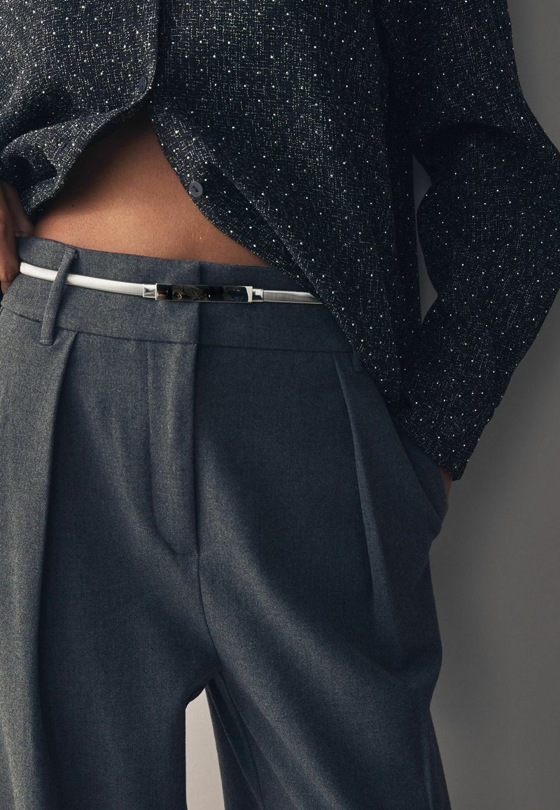 Graue maßgeschneiderte Hose mit Falten, kombiniert mit einem strukturierten dunklen Hemd, das einen subtilen metallischen Faden aufweist. Ein weißer Gürtel betont die Taille.