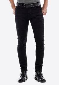 Zwarte slim-fit jeans met een gestructureerd oppervlak, vergezeld van een studded zwarte riem en gecombineerd met zwarte veterschoenen.