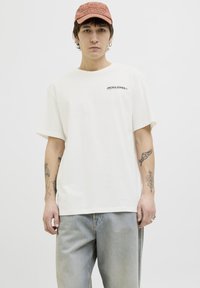 Witte t-shirt met korte mouwen met zwart "JACK & JONES" logo op de linkerborst, gecombineerd met lichtblauwe jeans, met zichtbare tatoeages op de armen.