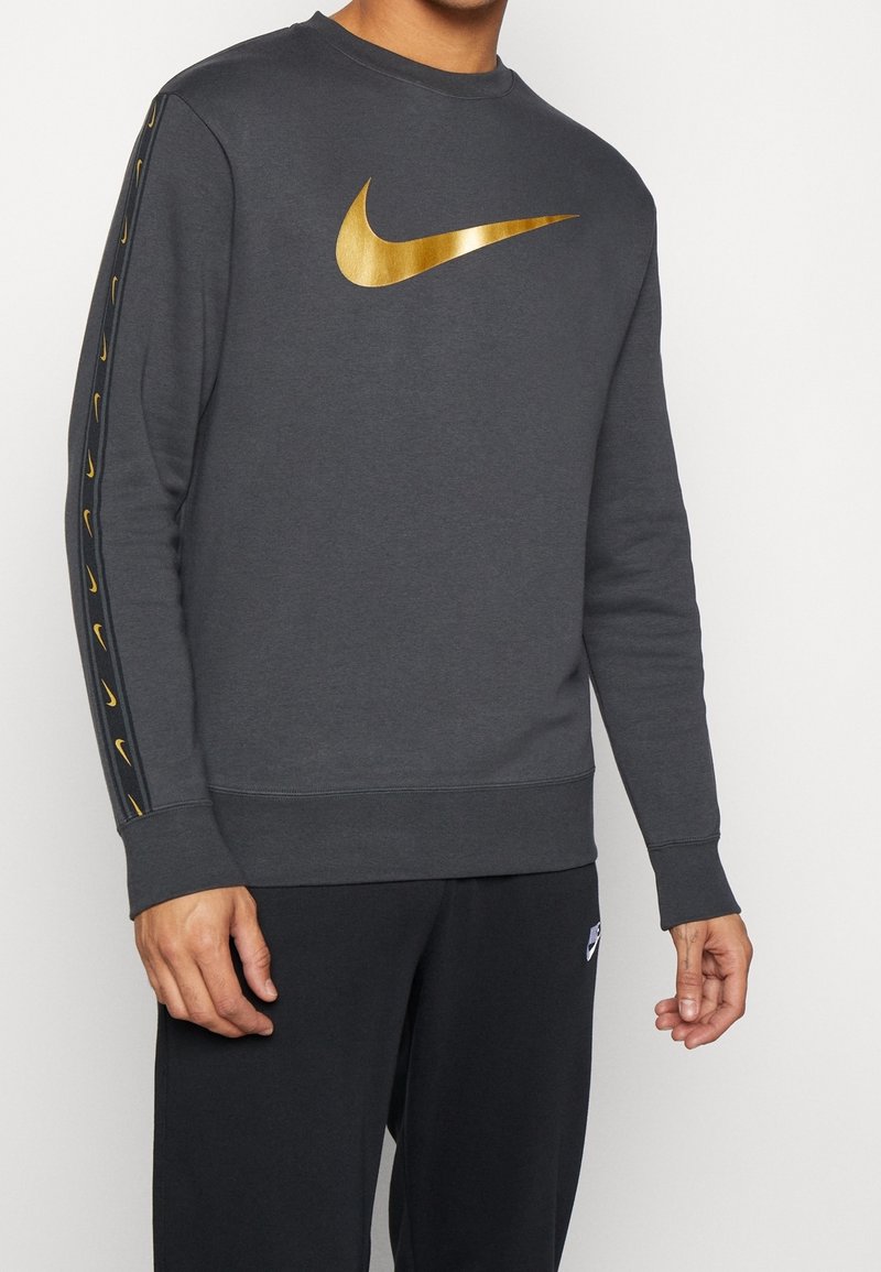 Tmavě šedá mikina s velkým zlatým logem Nike na hrudi, se zlatými detaily po stranách rukávů a žebrovanými manžetami.