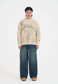 Beige gerippter Strickpullover mit grafischen Designs, kombiniert mit weit geschnittenen blauen Jeans und braunen Clogs. Weißer Hintergrund, neutrales Licht.