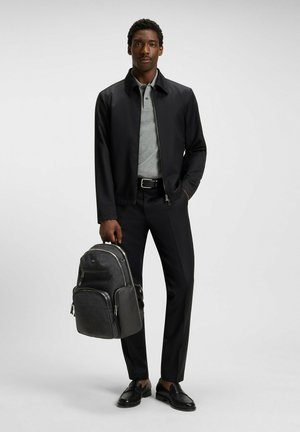 Veste zippée noire sur un polo gris, associée à un pantalon noir. Tenant un sac à dos gris et noir avec des accessoires en argent, mocassins noirs.